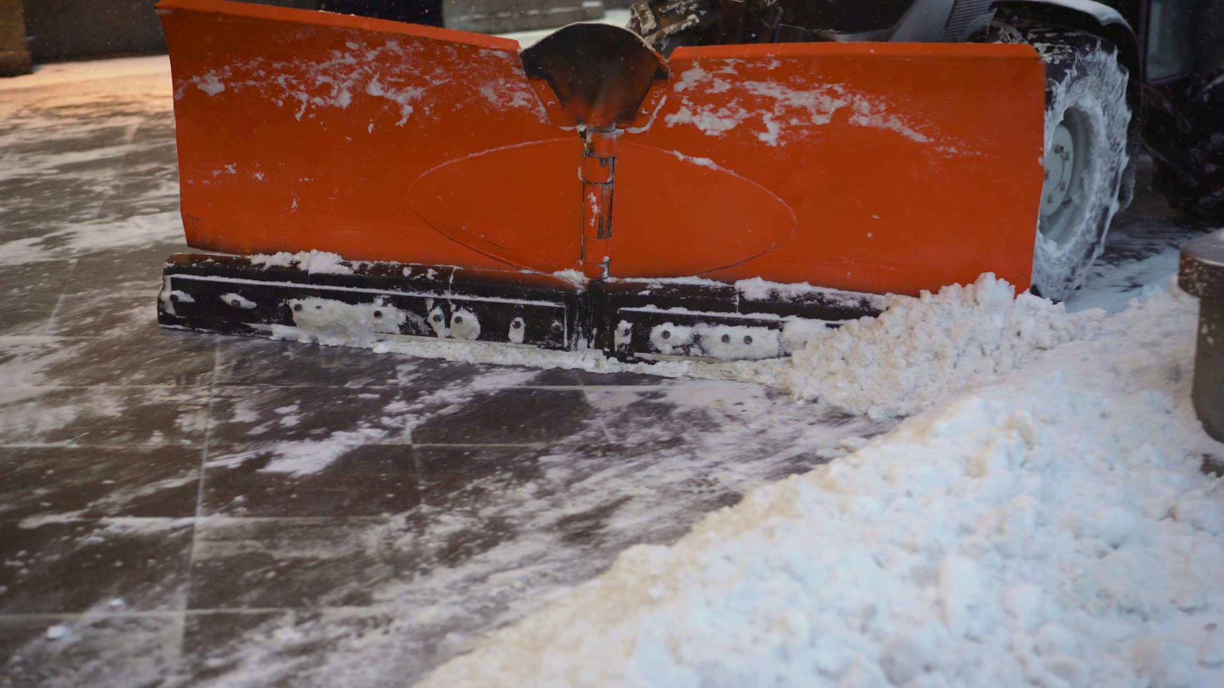 Orange snowplow clearing snow from tiled pavement